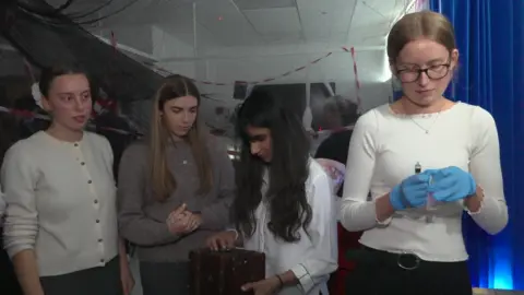 Four medical students, all women, are grouped together. One, with long dark hair, is looking at a box that needs unlocking. A fourth student, with straight blond hair tied back, has blue medical gloves on and a syringe in one hand. standing looking. One of the students, with dark blond hair tied back in a cream scrunchie is looking over at her. The lighting is subdued.