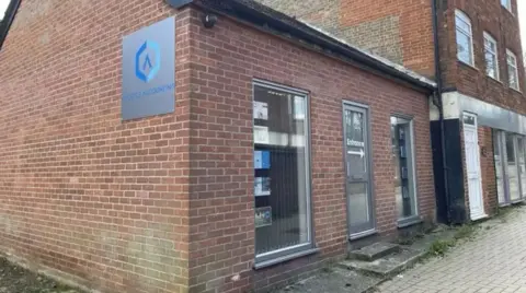 Front and side aspects of a single storey office building. The entrance and front windows are visible. The building is made out of brick and the pavement is visible out front.