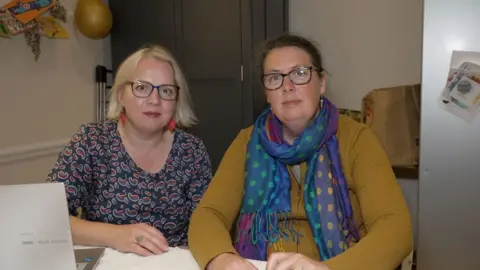 Iryna has short blonde hair and wears black rimmed glasses. She wears a dark blue patterned dress. Anna has dark hair tied up and wears brown rimmed glasses. She wears a yellow zip up top and blue shawl with polka dots. Both are sat at a desk with a laptop.