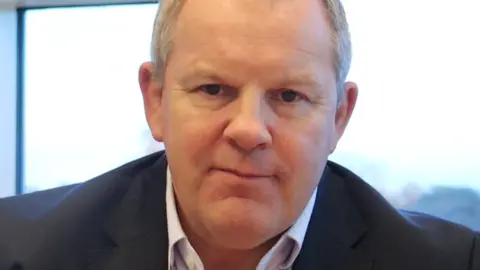 A picture of Steve McManus's head. He has short grey hair and is wearing a black jacket and white shirt. 