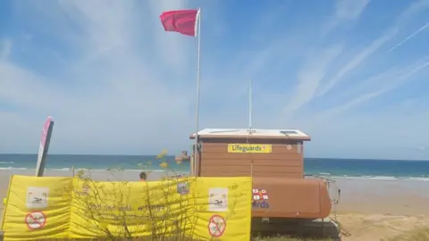 A red flag is flown above a brown, wooden lifeguard hut. It's overlooking a beach with the tide out and golden sand exposed. In the foreground, a yellow banner shows the rules around lifeguard flags.
