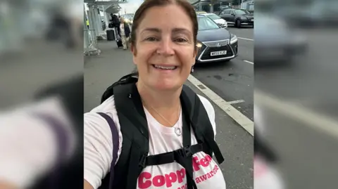 Rachael Kershaw A woman with brown hair smiling at the camera and wearing a black rucksack and pink t-shirt that reads Coppafeel!