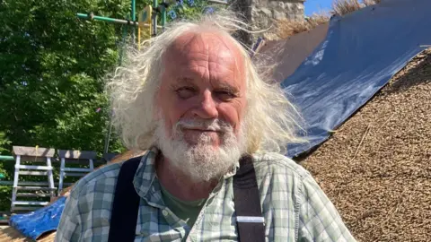 BBC Alan Jones wearing a checked green and white shirt with black braces. He is standing in front of a thatched roof.