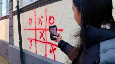Young at Heart A woman using her phone to capture graffiti on a wall, where two people appear to have played noughts and crosses with a red spray can. Crosses seems to have won, although the finishing move is drawn in what looks like pen - rather than the red paint used during the rest of the game.