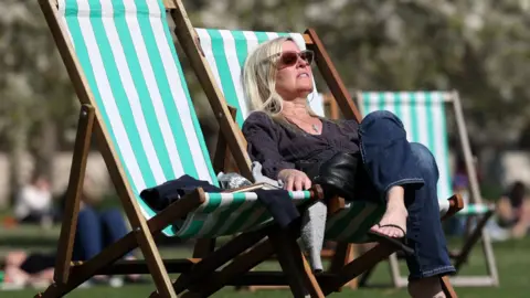 EPA-EFE/REX/Shutterstock A person relaxes in a striped deckchair on a sunny day, wearing sunglasses while sitting on grass in a park.