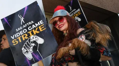 A costumed performer wears a fake fur coat and red-tinted sunglasses as she carries a placard with the words "Sag-Aftra video game strike" and a picture of a hand clutching a gamepad printed on it.