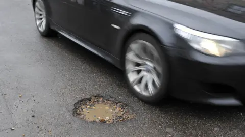 A black car driving past a water-filled pothole in the middle of a road
