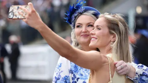 PA Media A woman in a yellow dress takes a selfie with her phone with a woman in a blue and white floral dress and a blue hat