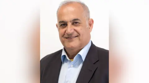 Somerset Council Habib Farbahi. He has short grey hair and is wearing a light blue shirt and a black suit jacket. He is looking at the camera and smiling.