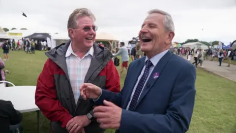 Two men chatting and laughing. The man on the left is wearing glasses and a red and grey jacket over a chequered shirt. The man on the right is wearing a blue suit and a tie. Behind them are people at a country show.
