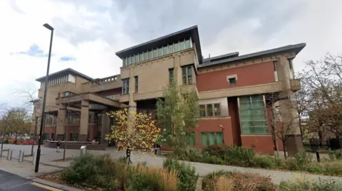 Google A modern-looking brown and red building with columns at the entrance and a sign saying Sheffield Crown Court. There are greenish high windows on the ground floor and rows of windows on the floor above. In front of the building is a pavement and greenery, including small trees and bushes.