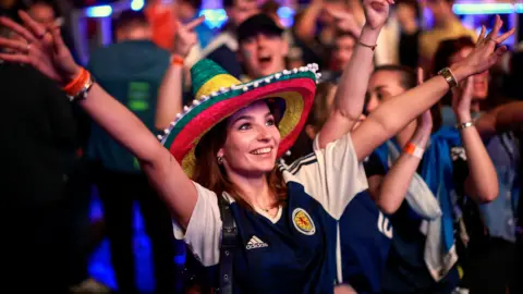 Getty Images A female fan wearing a colourful hat and a Scotland strip celebrates with her arms outstretched in the OVO Hydro. Rows of supporters can be seen in the background.