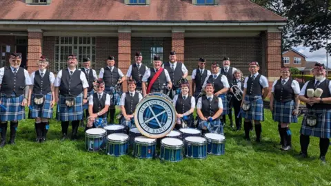 Newton Aycliffe Pipe Band The Newton Aycliffe Pipe Band are stood in a line with four members kneeling in front of them. A pile of drums are in front of them with one reading 'Newton Aycliffe Pipe Band' in writing curving around the drum. The band members are wearing blue kilts and formal uniforms.