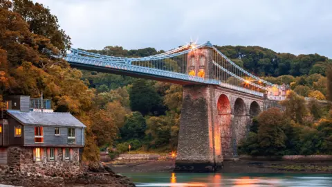 Menai Suspension Bridge, Anglesey, Wales Menai Bridge from The Belgian Promenade. A house can be seen on the left hand side under the bridge. It's dawn and lights illuminate the bridge. 