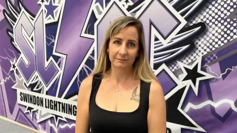 Swindon Lightning Cheerleading club director Millie Fannin pictured with long blonde hair and a black vest top standing in front of a purple, black and white back drop with the Swindon Lightning Logo on it.