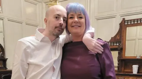 Roisin Wilshaw A man and a woman are looking at the camera - the man, who is bald with light facal hair and wearing a white shirt, is on the left. The woman, whose hair is dyed blue and violet, is on the right. She is wearing a burgundy top. In the background are white walls. 