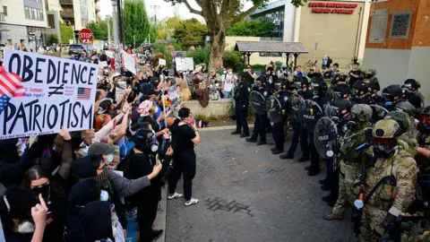 Getty Images Protesters stand with signs - some wearing masks - stand in a line facing off with authorities in Portland, Oregon. 
