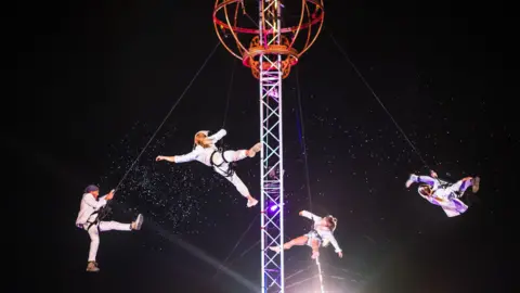 Four acrobats in white outfits hang from a large metal structure in the dark