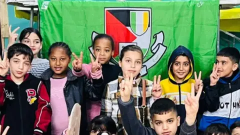 A group of young children from GAA Palestine pose alongside a green flag with the clubs logo