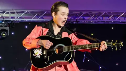 Cooper is on stage playing guitar and singing. He is wearing an orange satin suit and a long lapelled black shirt that has gold sequins on it. His hair is slicked back. 