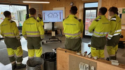 Scottish Power training centre, Cumbernauld - Several trainees wearing high‑visibility “ELECTRICITY” uniforms stand inside a training room, watching an instructor present a wiring diagram on a large screen.