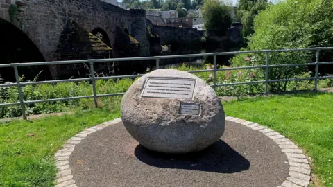 A large stone with a metal panel on it sits on tarmac beside a grassy area and a gate next to a bridge over the River Nith