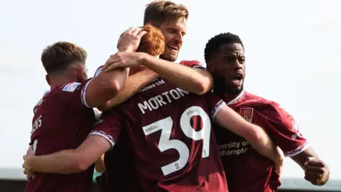 PA Media Northampton Town's Callum Morton celebrates with Sam Hoskins, Jon Guthrie and Luke Mbete during a League One match at Sixfields Stadium, Northampton, in August. They are wearing a claret kit and Morton and Mbete have their mouths open while celebrating. The other two have their backs to the camera.