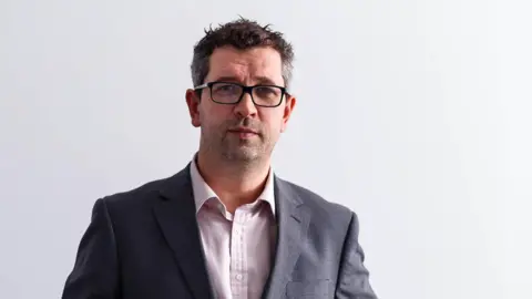 GWR A professional headshot of Dan Panes. He stands against a white backdrop and wears a grey suit jacket and a white shirt. He has short dark brown hair and wears black rimmed glasses.