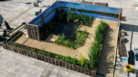 WWT An aerial view of an 8x8 installation with decking, grass, and water in Millennium Square in Bristol. There are construction workers each side of the installation.