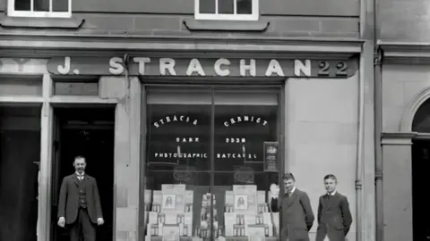 Three men in suits outside a shop called J.Strachan