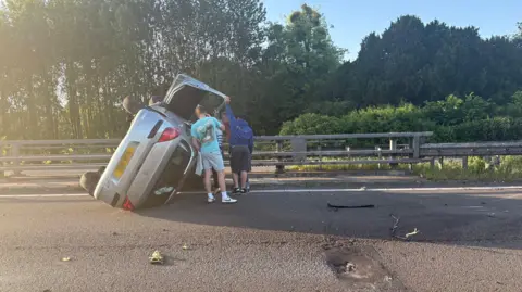 Three or four people stand beside a silver car on its side. The people hold open the door.