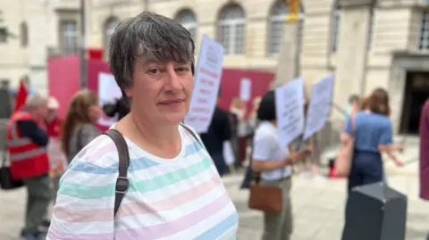 A woman wearing a horizontal striped top in green, white, pink and blue is standing facing the camera with other parent campaigners behind her. In the background is Leeds Civic Hall.