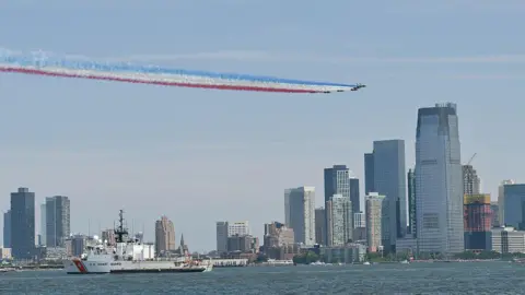 Aircraft trail red, white and blue smoke above waterfront skyline, passing US Coast Guard vessel