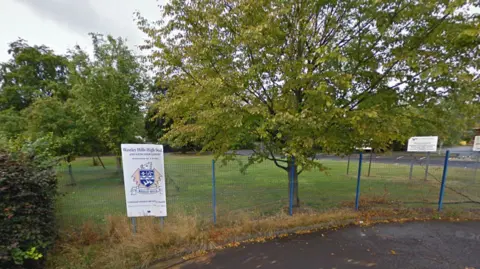 Google A photo of a school sign, white with a crest on it and reading Waseley Hills High School, on a blue security fence with trees on grass behind. 
