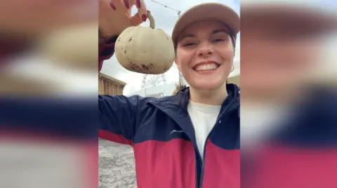 Jess⁤ Warren/BBC ‍Jess Warren in a blue and burgundy‍ raincoat and beige ⁤baseball cap. She holds a white pumpkin next to her head and smiles at the‍ camera.