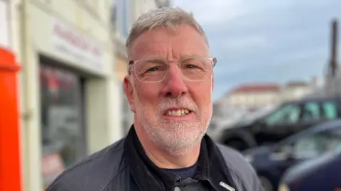 A man called Adrian Gabriel smiles at the camera. He has short white hairt and a trimmed white beard covering his face. He has clear-rimmed square glasses on. He is stood outside a parade of shops and parked cars, which are blurred behind him.