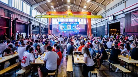 PROSPECT BUILDING A crowd of England fans sit around pub tables inside a large warehouse-style building 