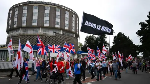 A group of protesters, many of whom are waving Union and St George's flags, march down a road. In the background The Roundhouse Hotel can be seen. 