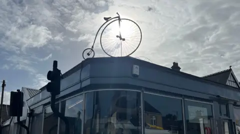 John Devine/BBC A model penny farthing bicycle with the sun behind it sits on top of a building, about 5m (16ft) up in the air. Traffic lights are partly visible and reflections in the glass windows show houses and cars.