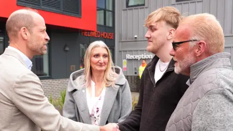 PA Media Prince William shakes the hand of a man with blond hair, a beard and wearing a black zip-up top and white-shirt. Looking on are a woman with long blonde hair and a man with short blond hair, sunglasses and a beard. Both are wearing black grey jackets. 