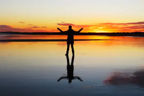 Alex Craig Sunset image showing a person with their arms outstretched on the beach, reflected in the sand