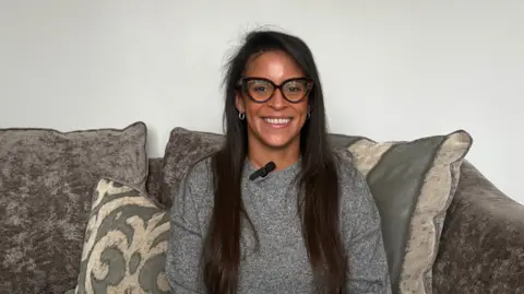The image shows a woman sat on a couch, smiling at the camera. There is a microphone clipped to the collar of her jumper and she is wearing glasses with black frames. There are cushions behind her and the wall of the room is white.