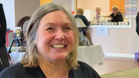 Paula Nelson has blond hair to her shoulders and is smiling, surrounded by items for sale at a coffee morning with ladies in the background serving hot drinks.