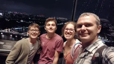 Toby Wells A selfie of a man with grey wearing a blue and white checked shirt. He stands beside a woman with glasses and coral t-shirt, and two young boys with brown hair. One is wearing a red t-shirt and another is wearing glasses with a grey t-shirt and green jacket.