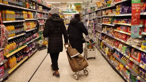 EPA/Shutterstock Customers holding a basket and wheeling a trolley bag shop walk down a supermarket aisle.