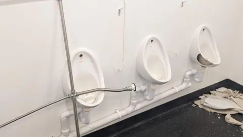 East Suffolk Council Three white urinals are on a white wall in a public toilet. The one at the far right is badly chipped and there is a pile of broken porcelain on the dark floor beneath it
