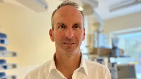 The image shows a slim, middle-aged man with a white shirt. He is looking at the camera with a neutral expression. He is standing in a clinical room in a hospital with lots of machines behind him. 