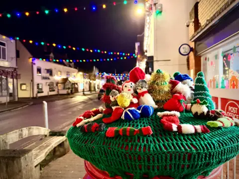 BBC Weather Watchers/Snaphappydave A green post box topper has a number of Christmas characters, trees and crackers knitted and sewn into its surface. The well-lit, night-time scene has Christmas lights strewn above the street in a zig-zag formation down the road.