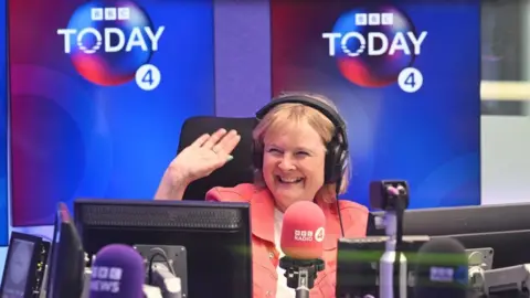 Jeff Overs/BBC Martha Kearney, wearing a pink jacket and white top, smiles and waves in a radio studio, surrounded by BBC Radio 4 branding