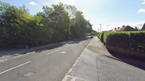 Google The junction of two roads. There are hedges either side of the road with a few rooftops visible in the distance.